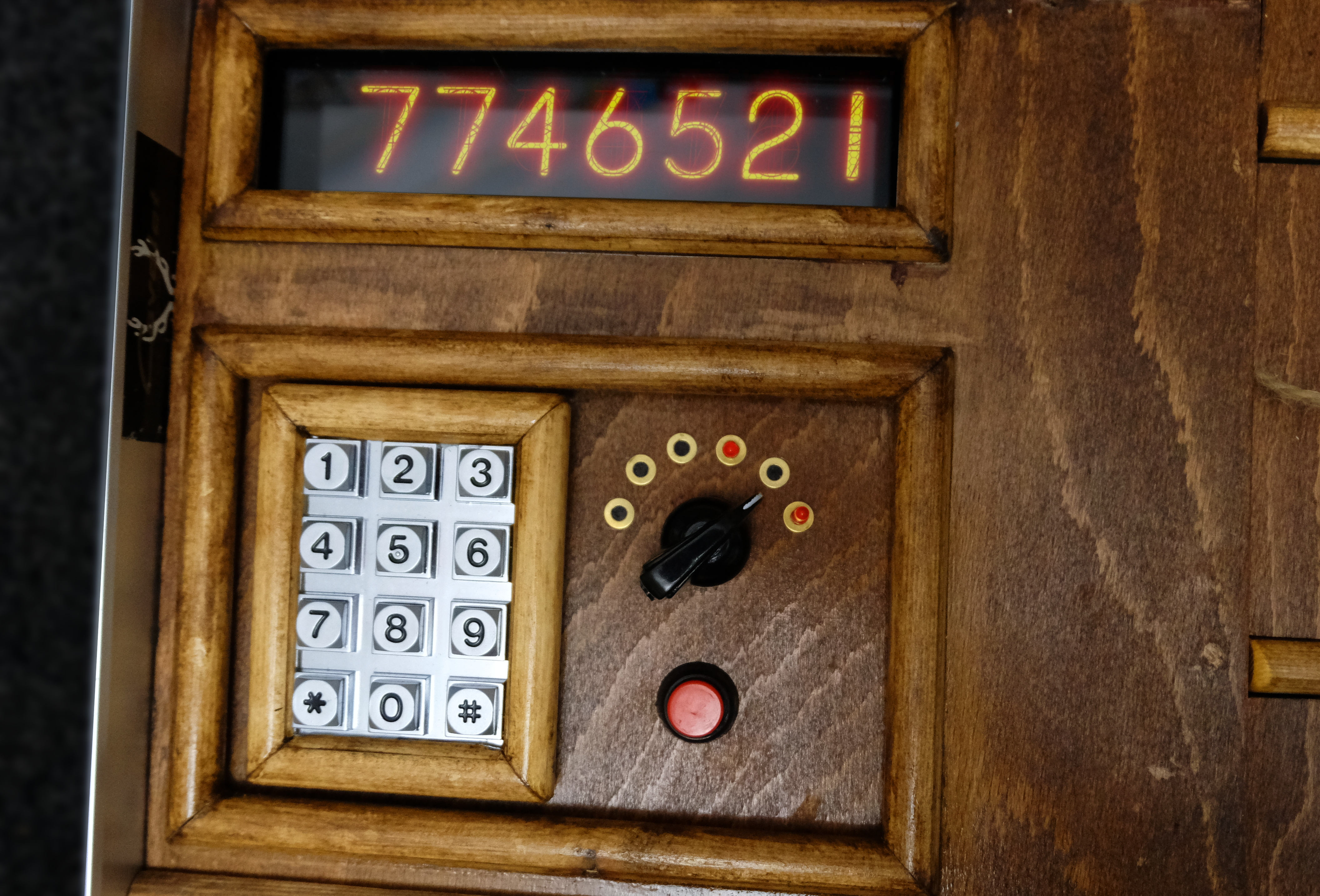 A wooden framed contraption has a narrow screen shows a series of illuminated digits, below that is a numerical keypad, a dial, and a button